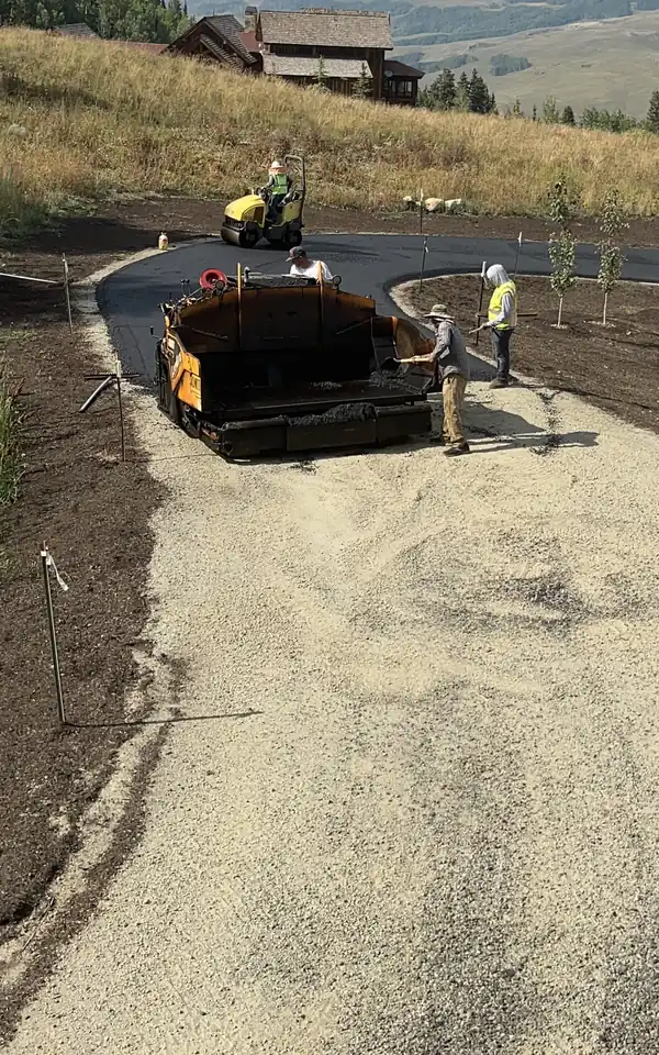 Paving crew laying a new asphalt driveway up to a mountain home, with high peaks and open meadows behind