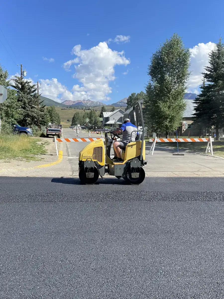Ride-on roller compacting fresh asphalt on a mountain-town street with peaks in the distance